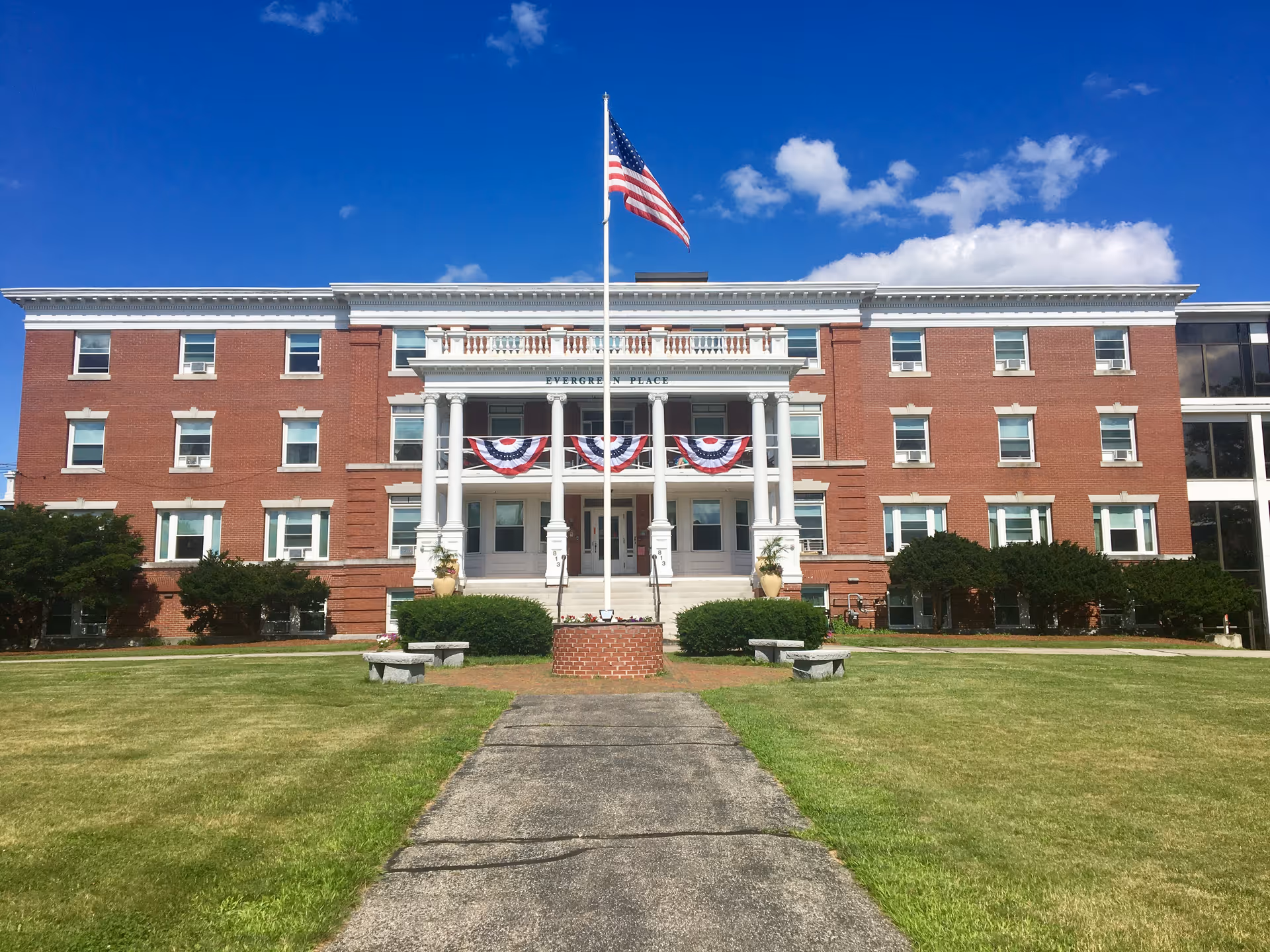 Brick Evergreen Place building with white columns, flagpole, bunting, and green lawn under blue sky.