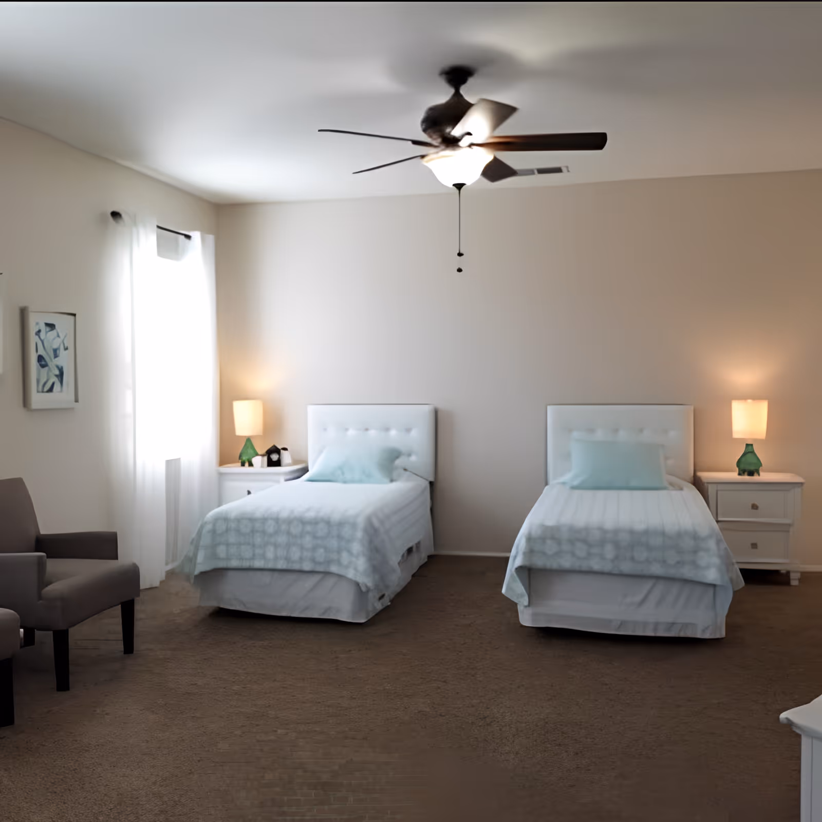 A bedroom with two single beds each with white tufted headboards, light blue pillows, and matching light blue patterned blankets. Between the beds are two white nightstands with green glass lamps and small decorative items. A ceiling fan with a light is centered above the beds. To the left, there is a window with sheer white curtains and a gray armchair nearby. The walls are painted beige and the floor is carpeted in brown.