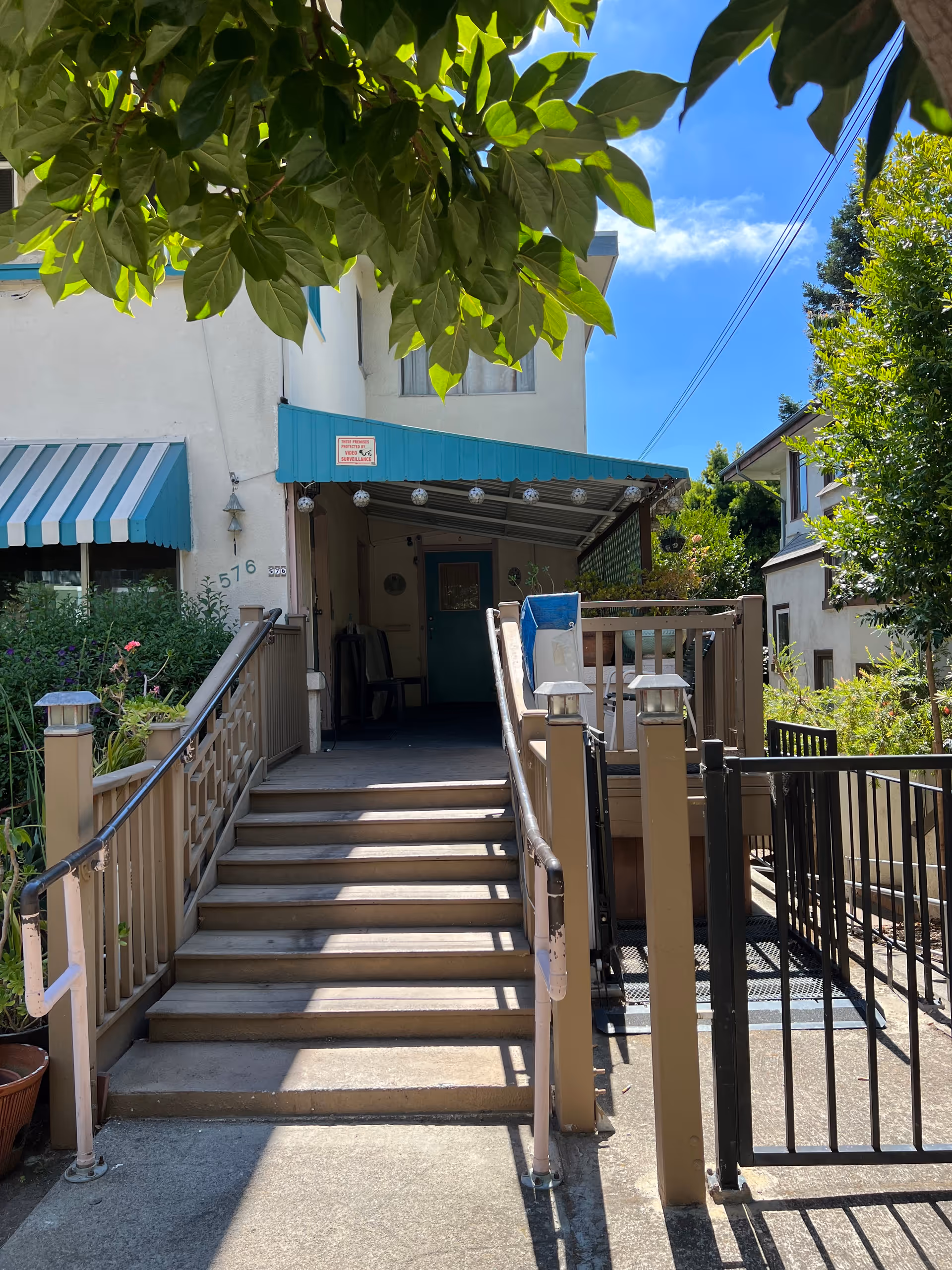 Entrance to a building with a staircase and a wheelchair ramp leading up to a covered porch area. The building has a white exterior with a blue awning above the door and windows. There are plants and greenery around the entrance, and a clear blue sky is visible above.