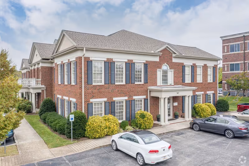 Brick senior living facility exterior with front entrance, windows, shrubs, and parked cars.