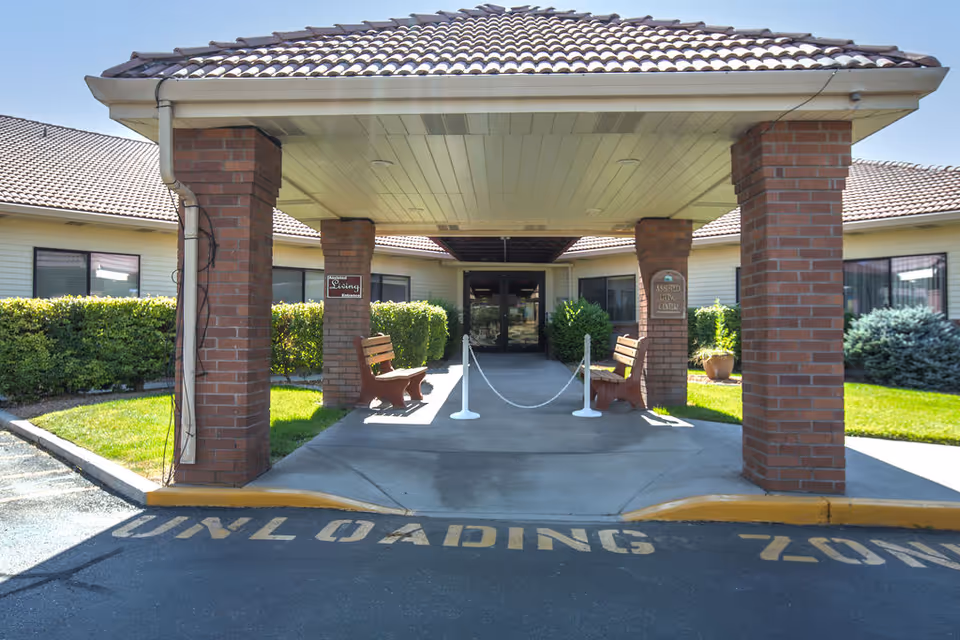 Covered driveway entrance with brick columns, benches, and a rope barrier leading to the front doors of a senior living facility.