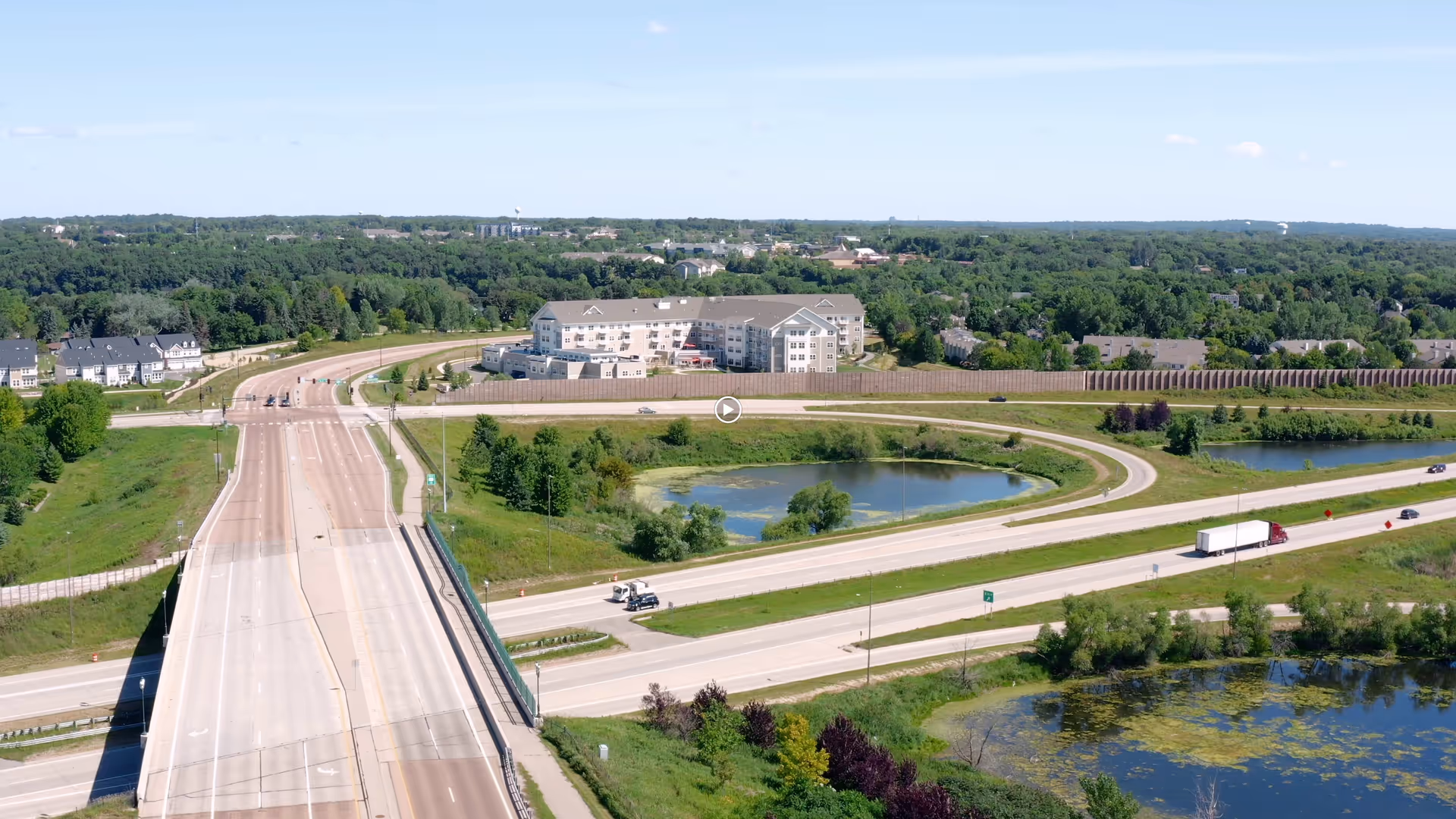Aerial view of a senior living facility named Riley Crossing Senior Living situated near a highway with surrounding greenery, ponds, and a road interchange.