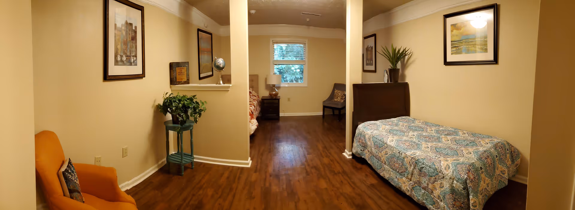 A cozy bedroom with two beds separated by a partial wall. The room has wooden flooring, beige walls, and framed artwork on the walls. There is an orange armchair with a decorative pillow on the left side, a small green plant on a stand, a nightstand with a lamp near the window, and a gray chair in the corner.