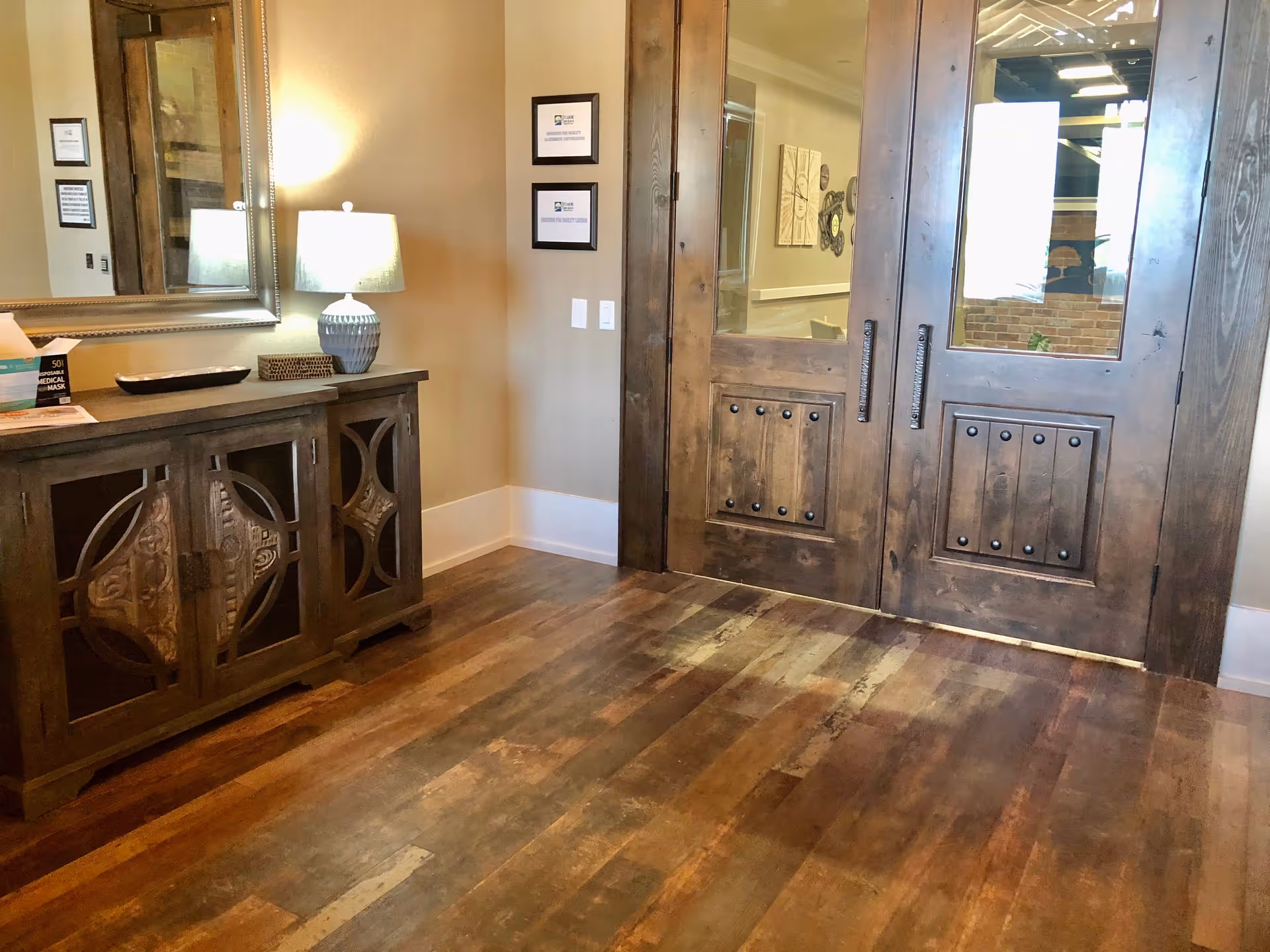 Interior view of a room with wooden double doors featuring glass panels. To the left, there is a wooden cabinet with decorative carved panels, topped with a table lamp, a mirror on the wall above, and some small items including a box of disposable medical masks. The floor is wooden with a rustic finish, and the walls are painted beige with white baseboards.