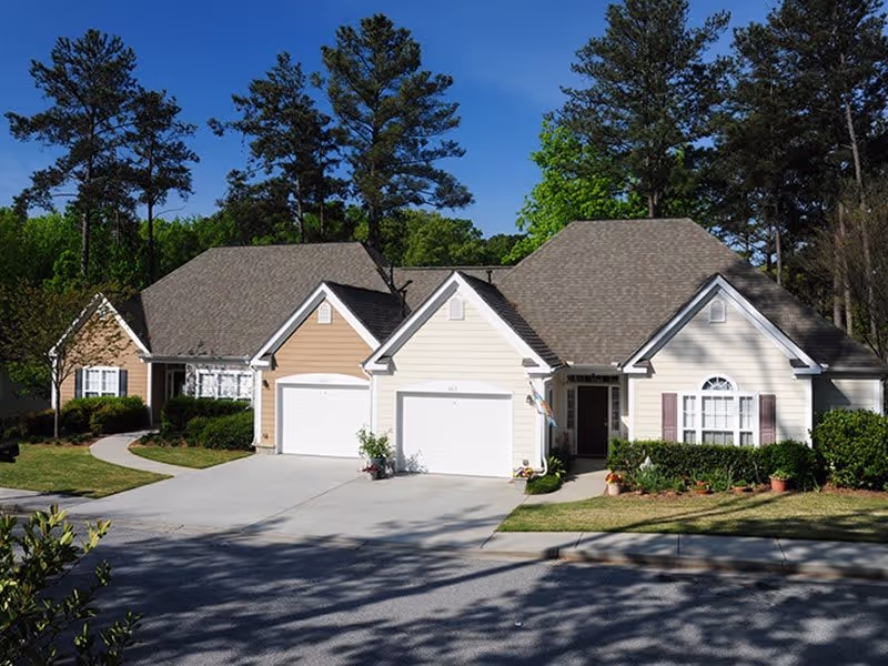 Two attached single-story homes with front garages, landscaped lawns, and tall pine trees behind them.