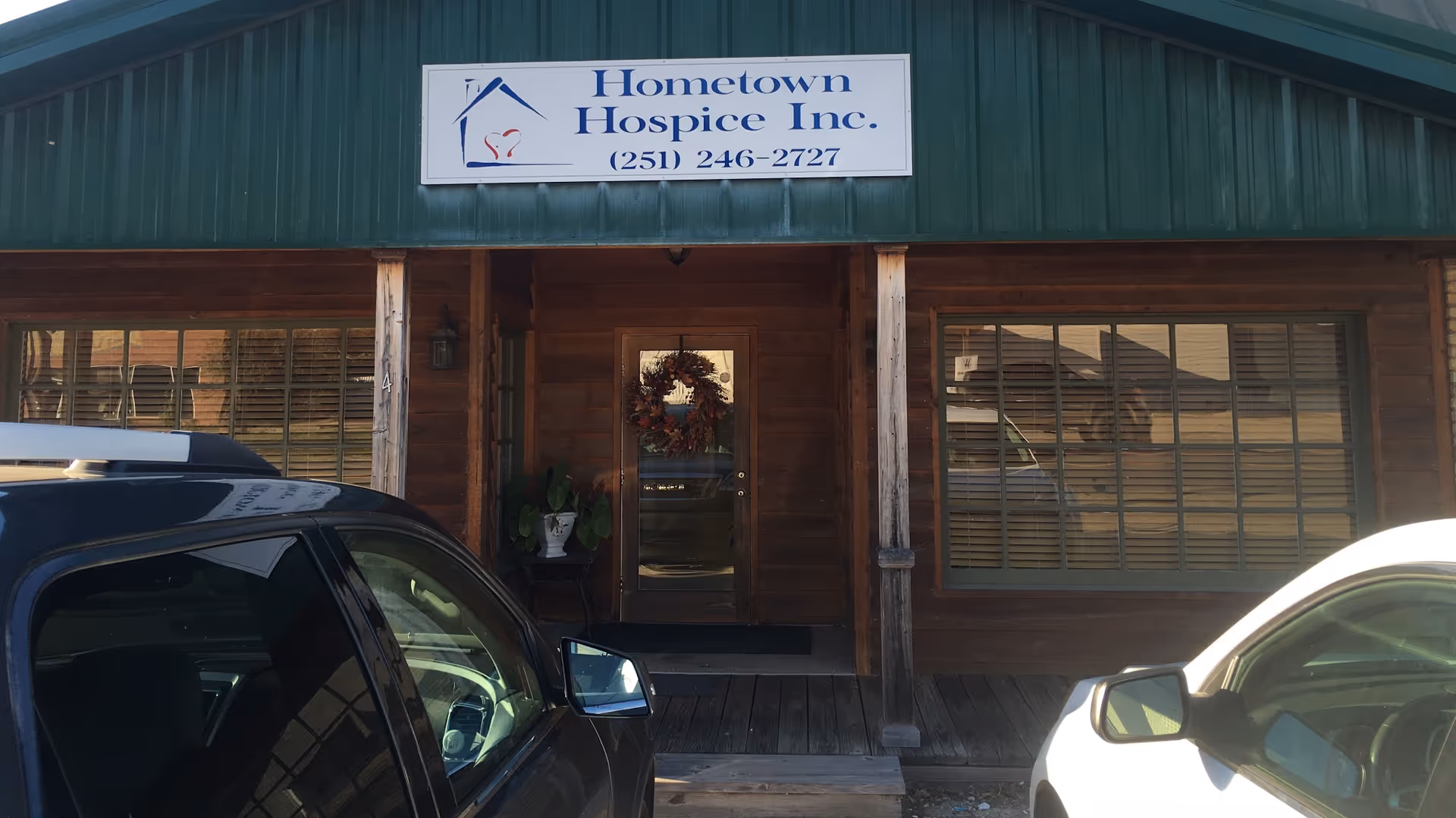 Wooden storefront with Hometown Hospice Inc. sign, wreath on door, and two parked cars.