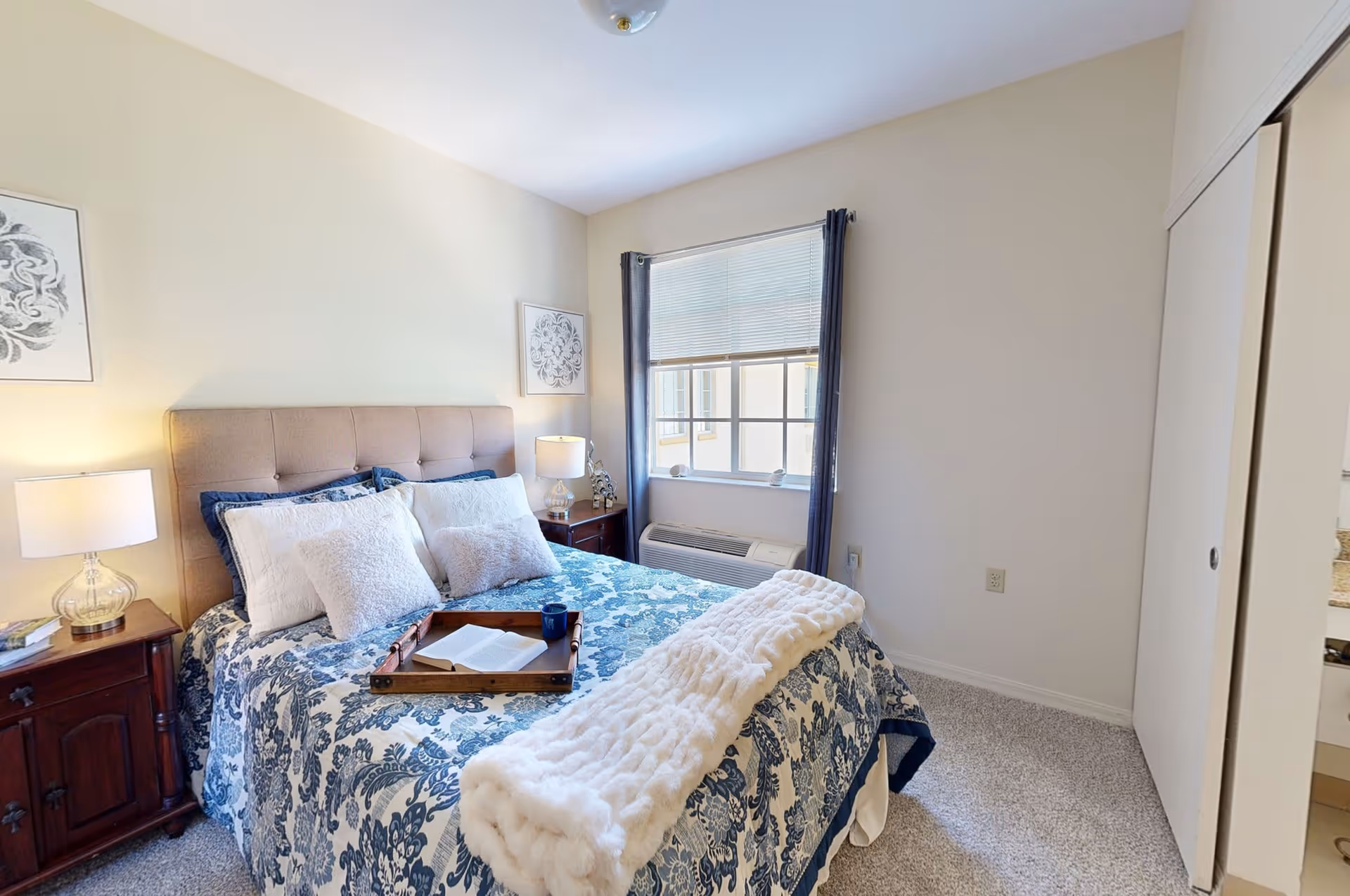 A cozy bedroom with a bed featuring a blue and white floral patterned comforter, multiple pillows, and a white throw blanket. There are two wooden nightstands with lamps on either side of the bed. A window with blinds and dark curtains is on the wall behind the bed. The room has light-colored walls and carpeted flooring.