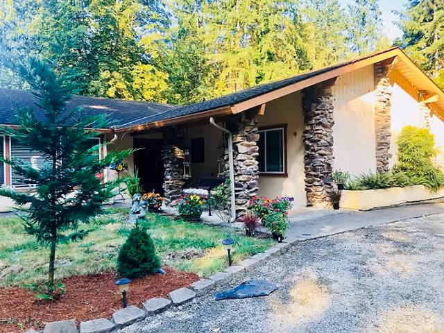 Exterior view of a single-story building with stone pillars and a sloped roof, surrounded by greenery and small landscaped garden beds with flowers and shrubs. A gravel driveway leads up to the entrance, and tall trees are visible in the background.