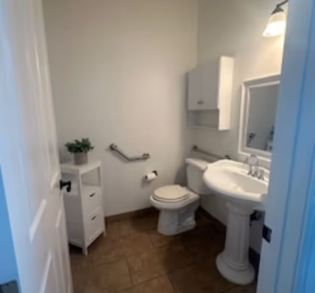 Small senior living bathroom with toilet, pedestal sink, mirror, and grab bars.