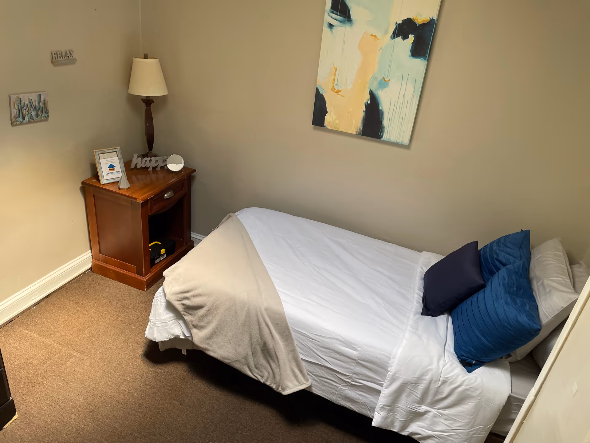 Small bedroom with a single bed, nightstand, lamp, and wall art in a neutral-toned room.
