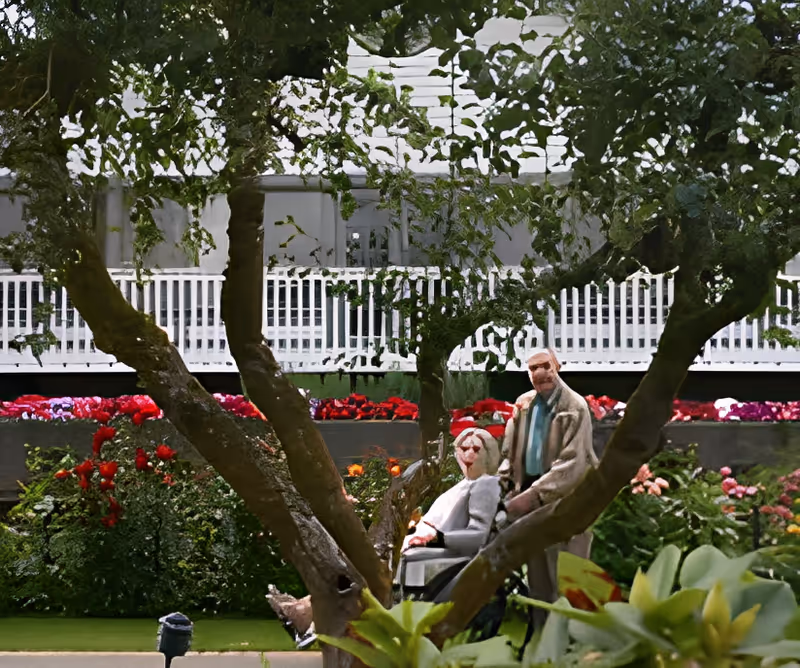 An elderly man standing behind an elderly woman in a wheelchair in a garden area with colorful flowers and greenery. In the background, there is a white building with a porch and railing.