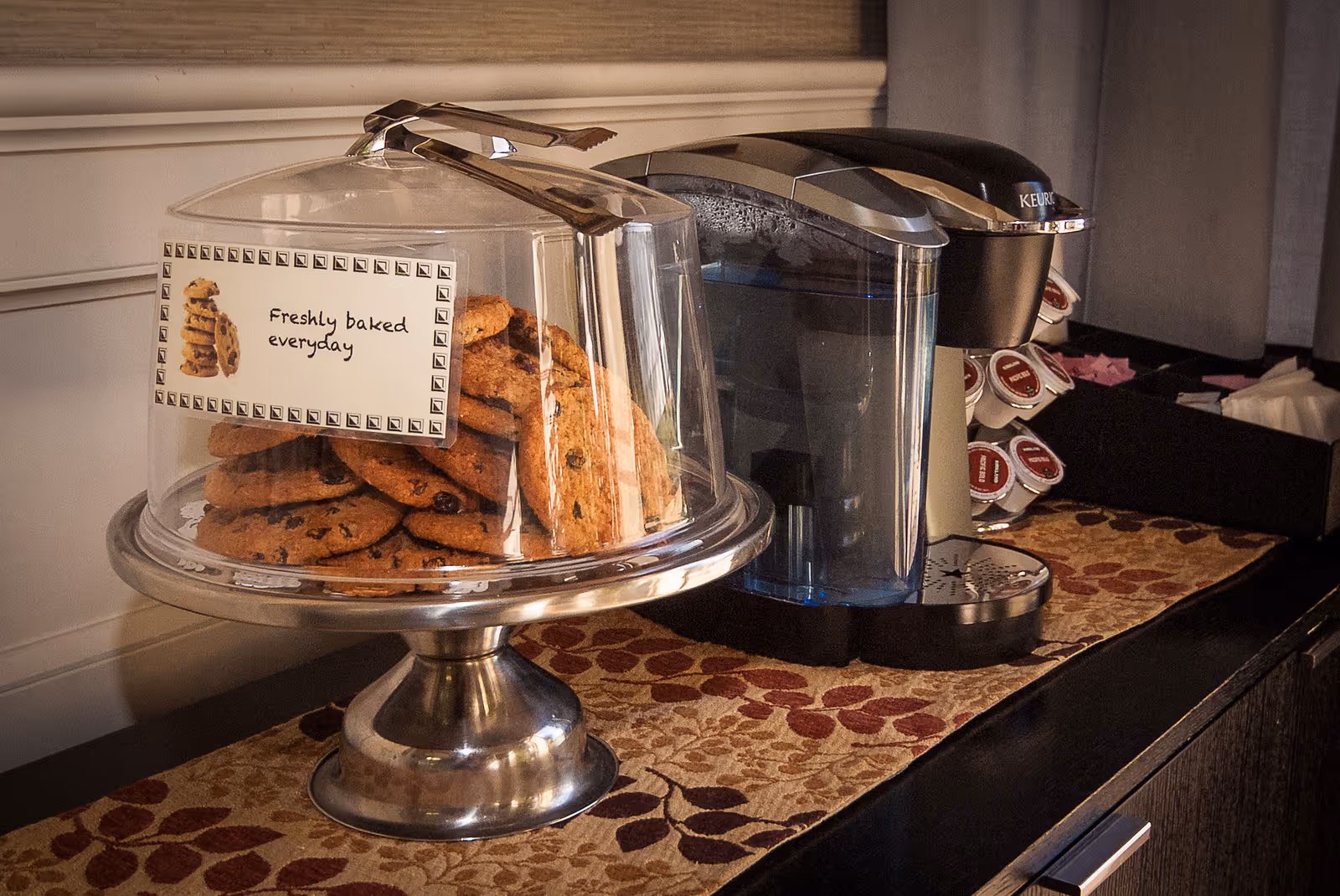 A glass-domed tray of cookies labeled "Freshly baked everyday" next to a Keurig coffee maker and K-Cup holder on a countertop.