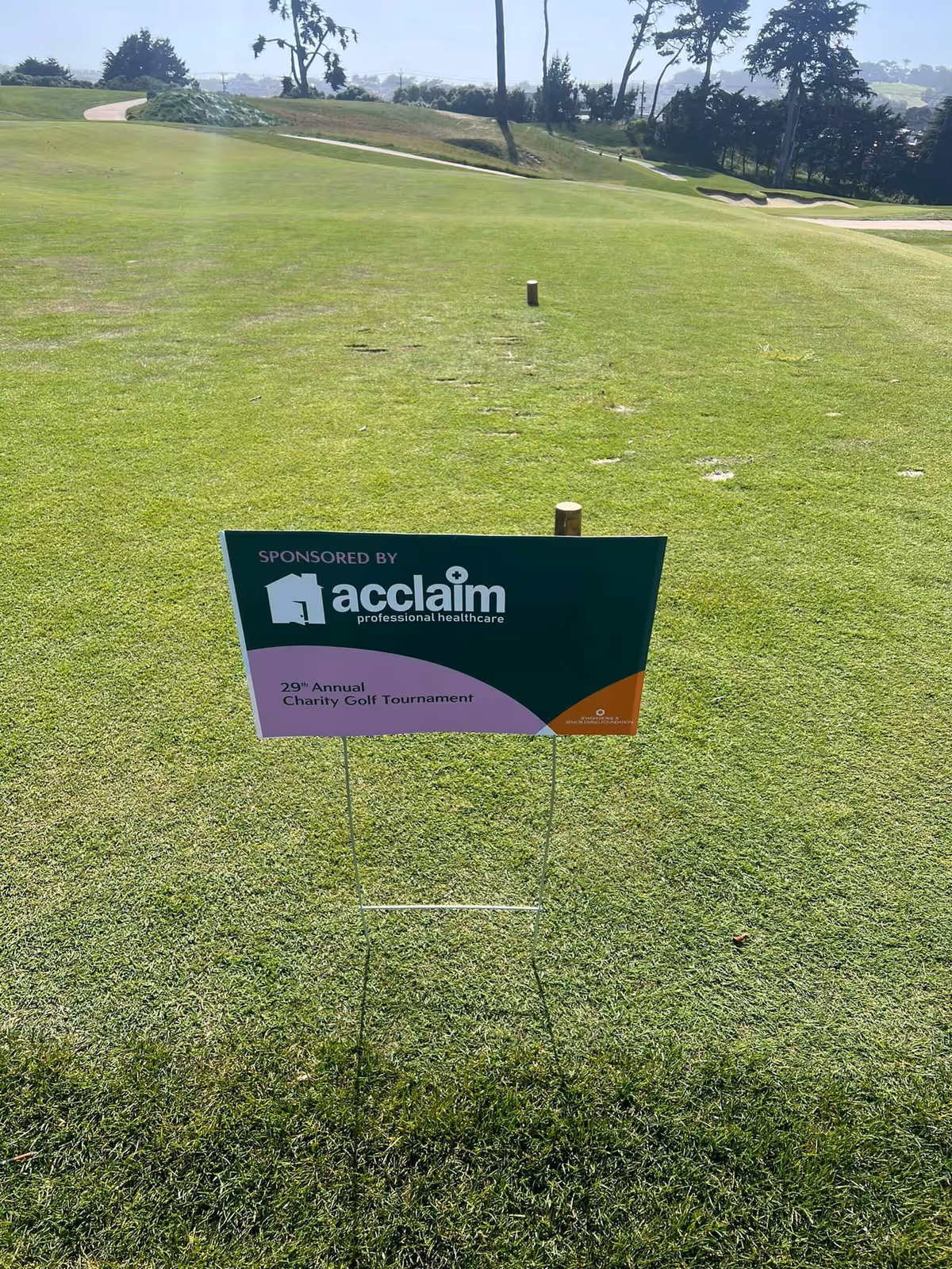 Sunlit golf course with Acclaim Home Care sponsor sign at charity tournament.