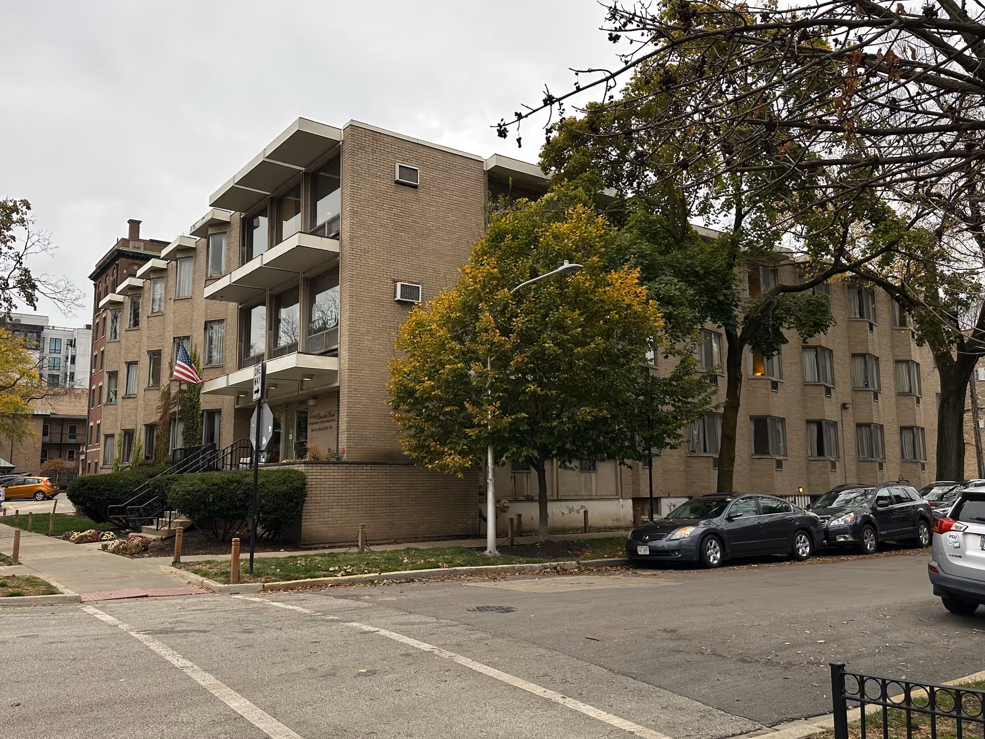 Exterior view of a multi-story brick senior living facility with balconies, trees, parked cars, and an American flag.