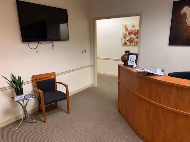 Reception area with curved wooden desk, mounted TV, chair, plant, and doorway.