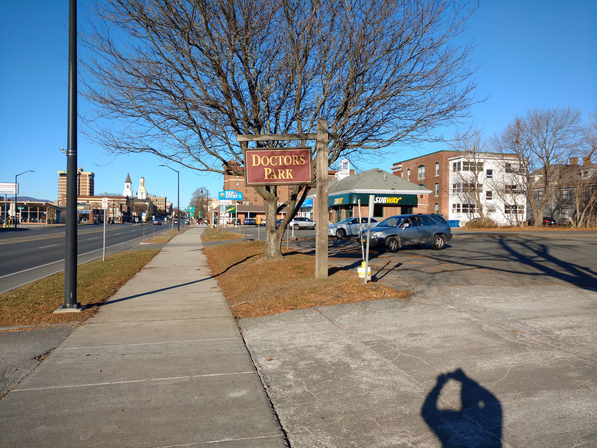 Wide street scene with a Doctors Park sign, leafless tree, sidewalk, and parked cars.