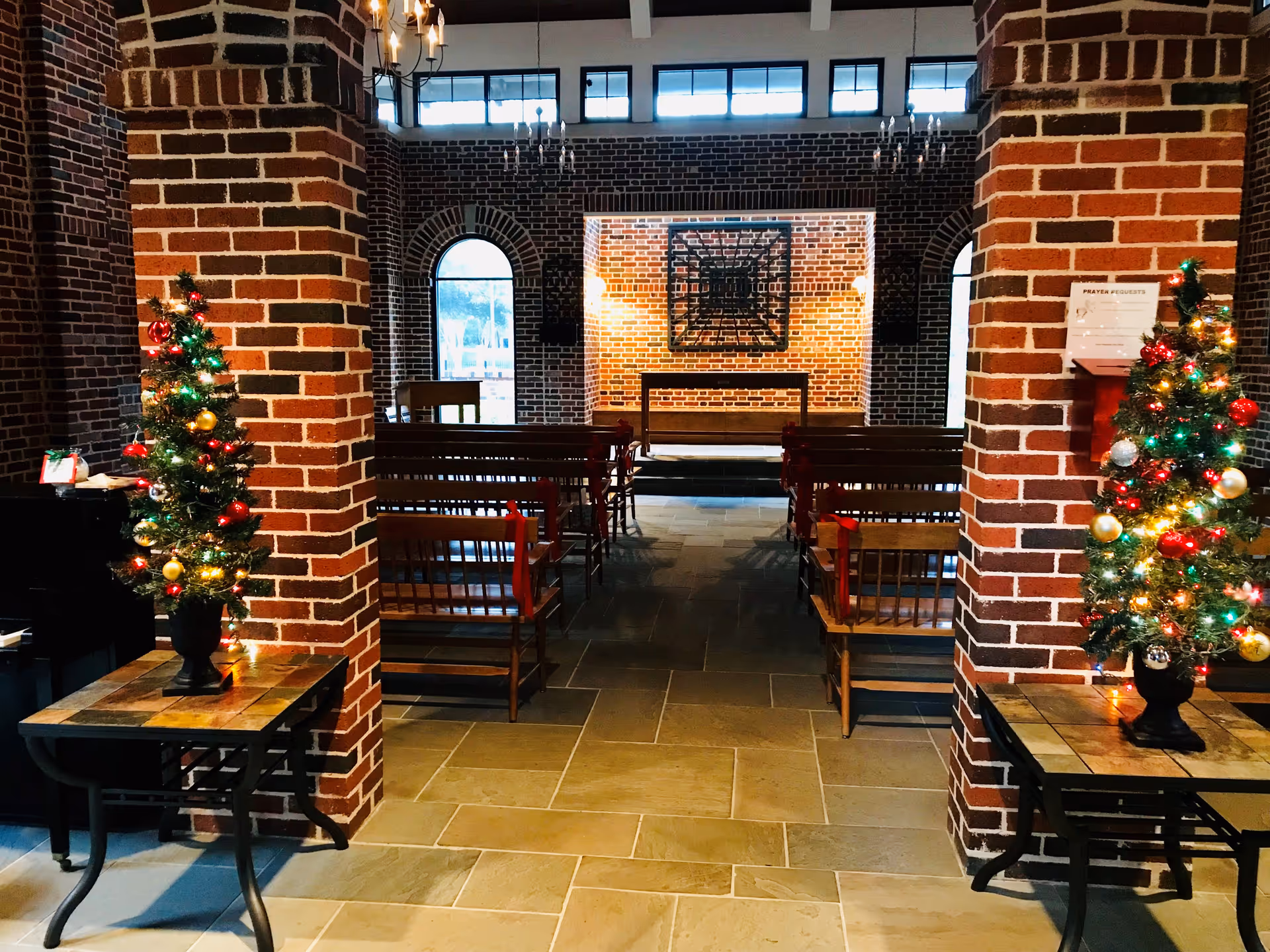Brick-walled chapel interior with pews, arches, and two Christmas trees near entrances.