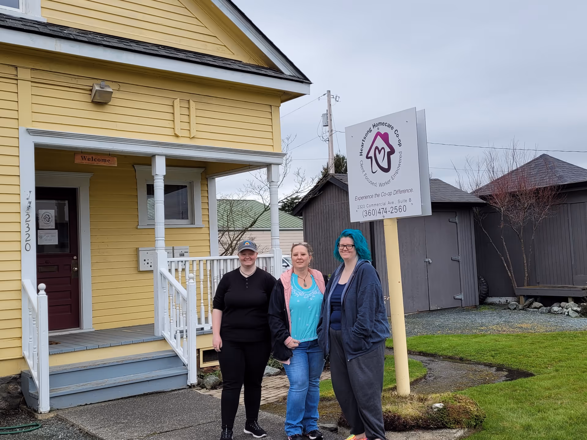 Three people stand in front of a yellow building with a Heartsong Homecare sign.