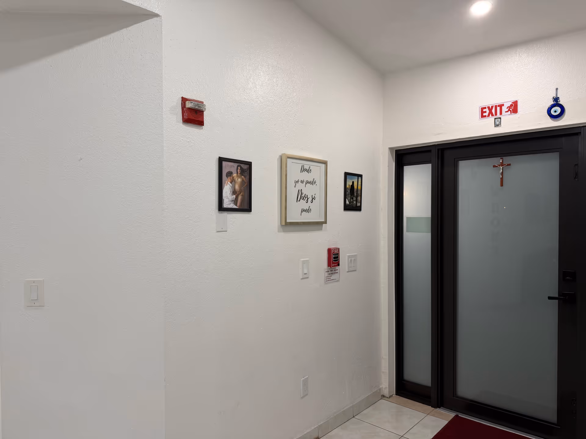 White hallway with wall art, frosted glass door, exit sign, and a cross.
