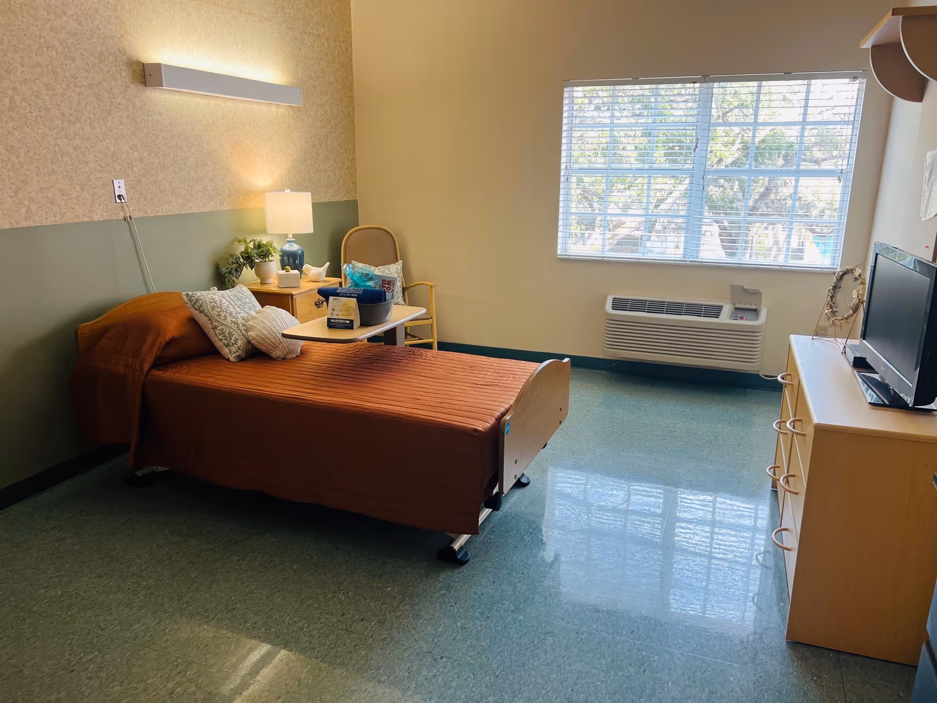 Single-bed patient room with bedside table, lamp, dresser, TV, and a large window.