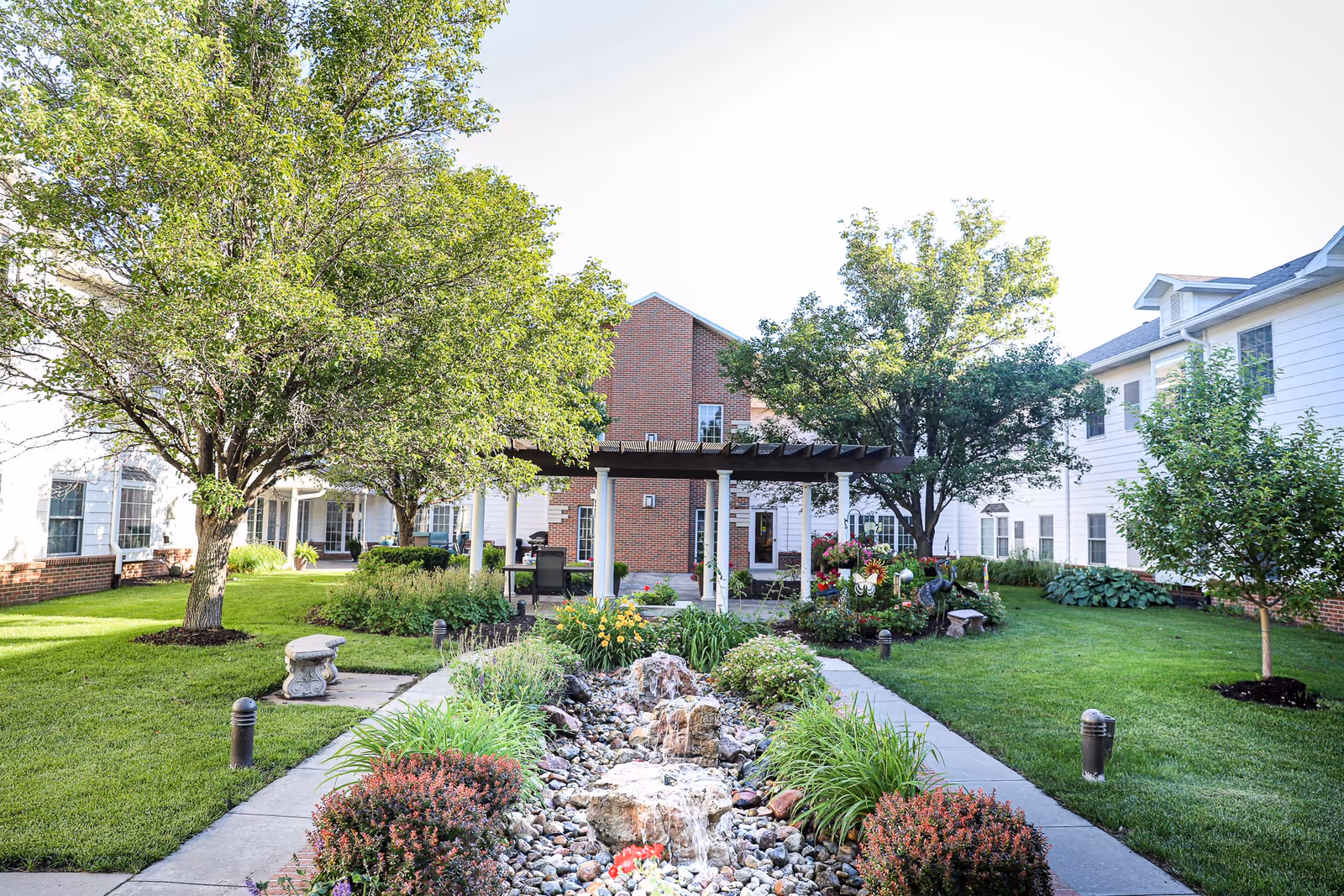 A landscaped outdoor garden area with a small rock water feature in the center, surrounded by green grass, bushes, and trees. There is a paved walkway on either side of the water feature leading to a pergola-covered seating area in front of a brick and white building.