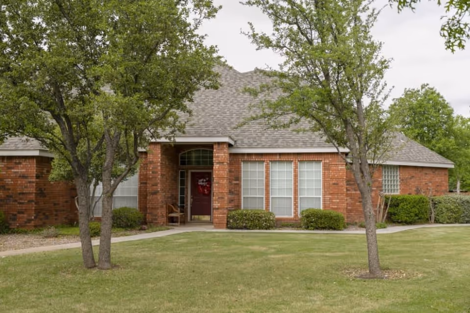 Single-story red-brick building facade with a lawn, trees, and a covered entrance with a red door.