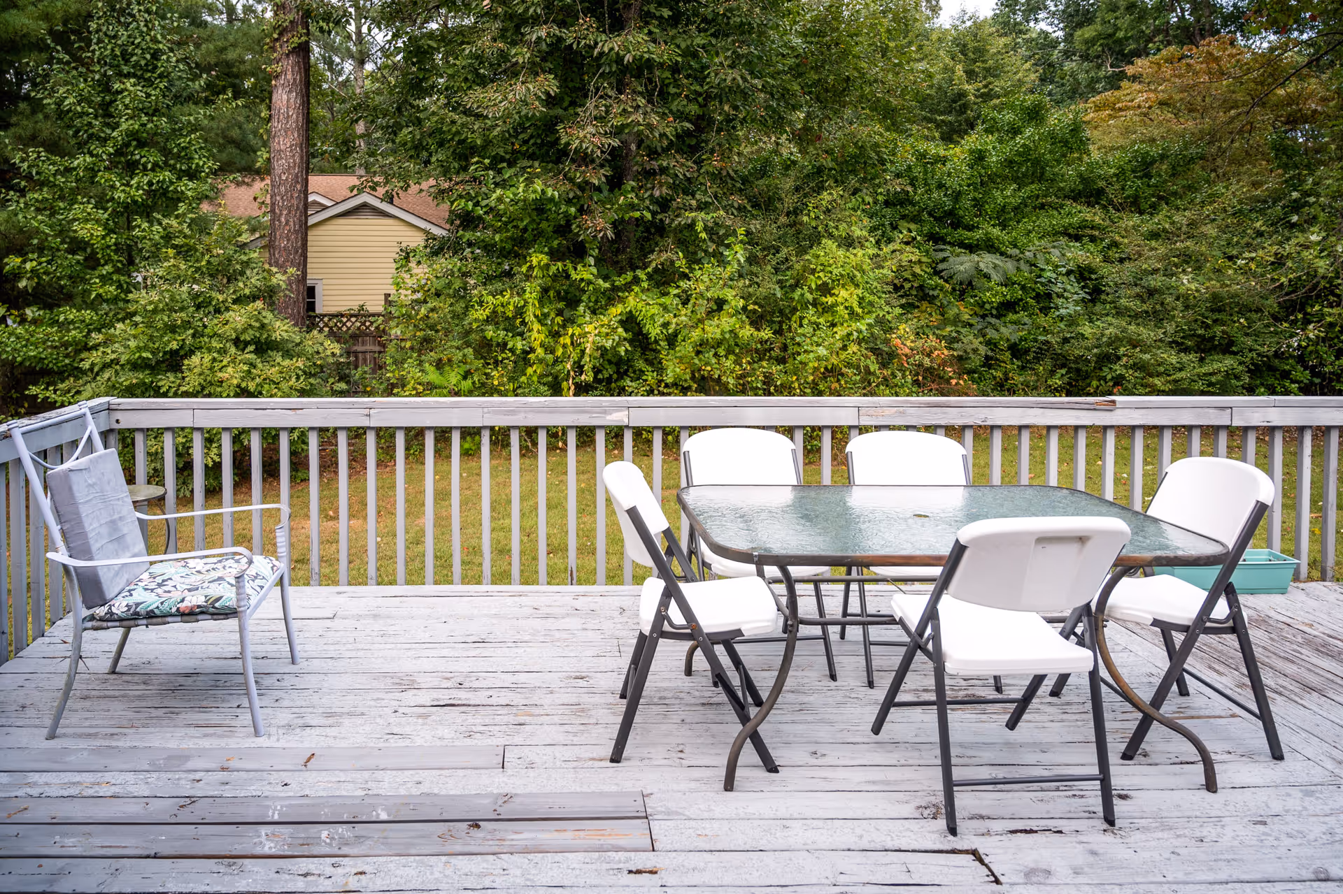 Outdoor deck with glass table, white chairs, and surrounding trees.