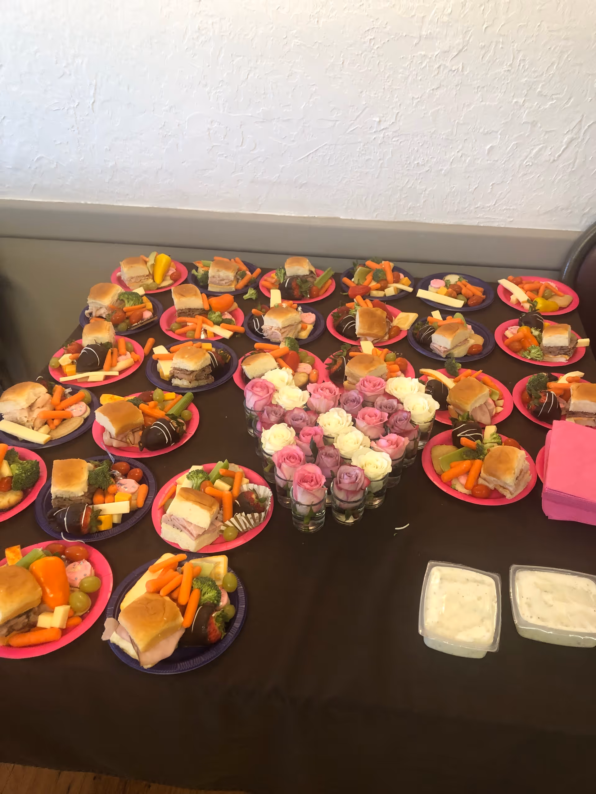 A table covered with a dark tablecloth displaying multiple plates of food, each containing a sandwich, baby carrots, broccoli, cheese sticks, grapes, cherry tomatoes, and a chocolate-covered strawberry. In the center of the table is a heart-shaped arrangement of pink and white roses in small glass vases. Two containers of white dipping sauce and a stack of pink napkins are also on the table.