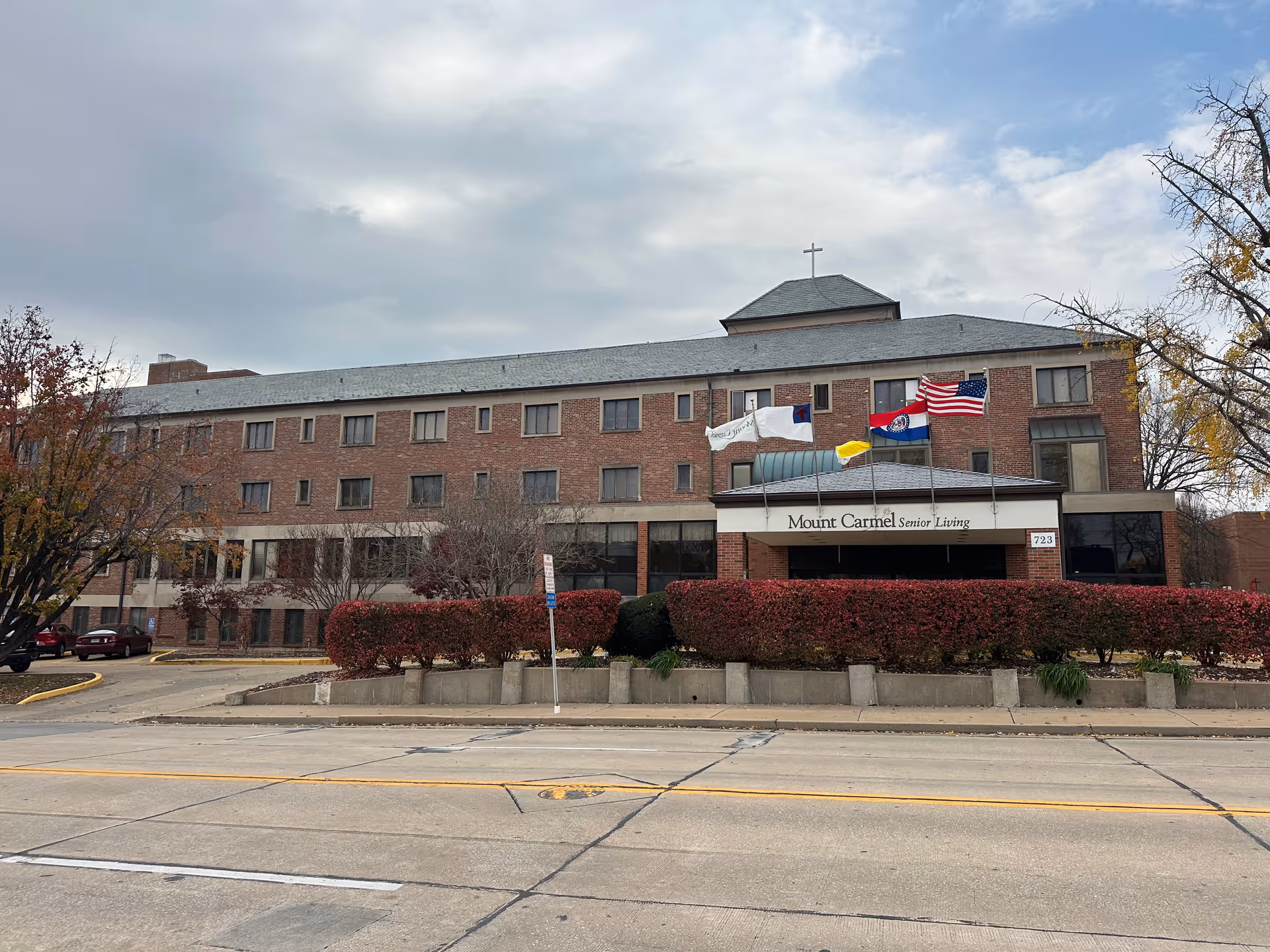 Exterior view of a three-story brick building with a sign that reads 'Mount Carmel Senior Living' above the entrance. There are four flags flying on poles in front of the building, and trimmed red bushes line the entrance area. The sky is partly cloudy.