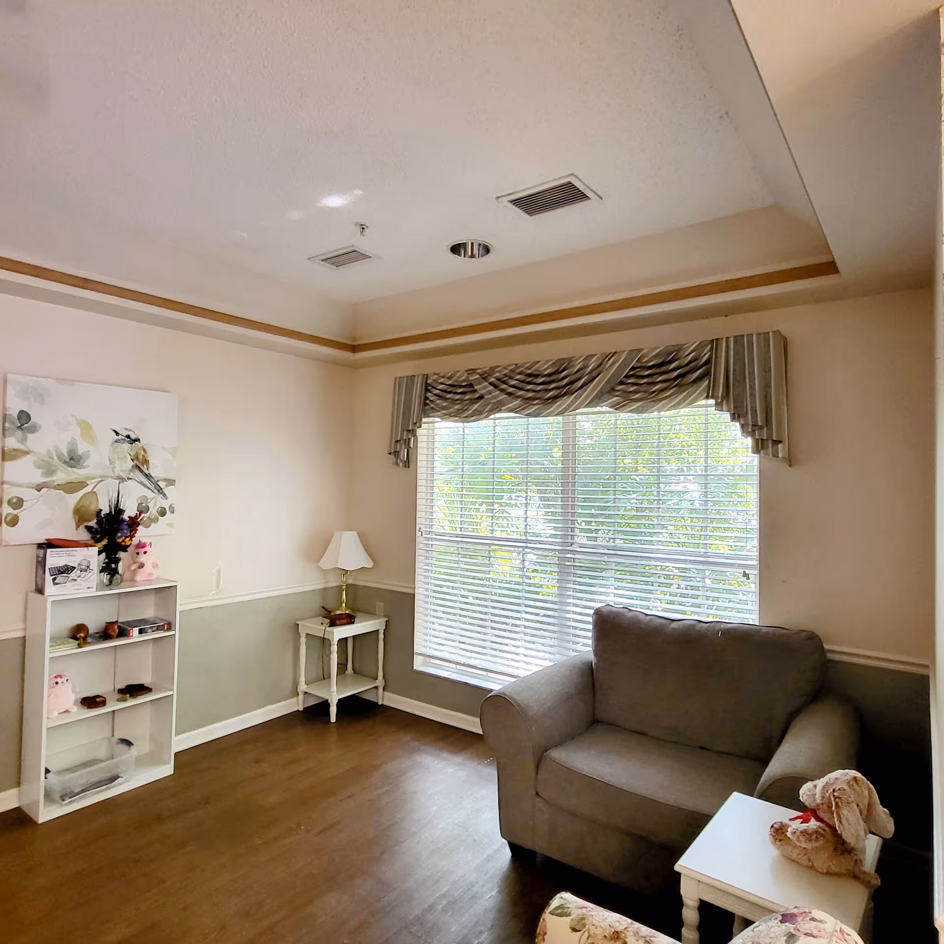 Cozy living room with grey couch, side tables, lamp, and large window with blinds.