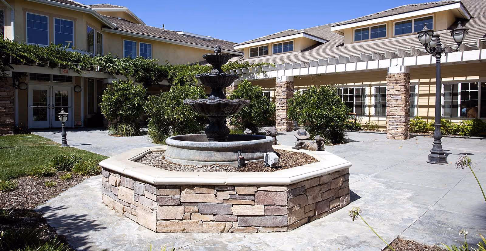 Stone three-tier fountain in a paved courtyard in front of a senior living building with a pergola, lamp posts, and landscaping.