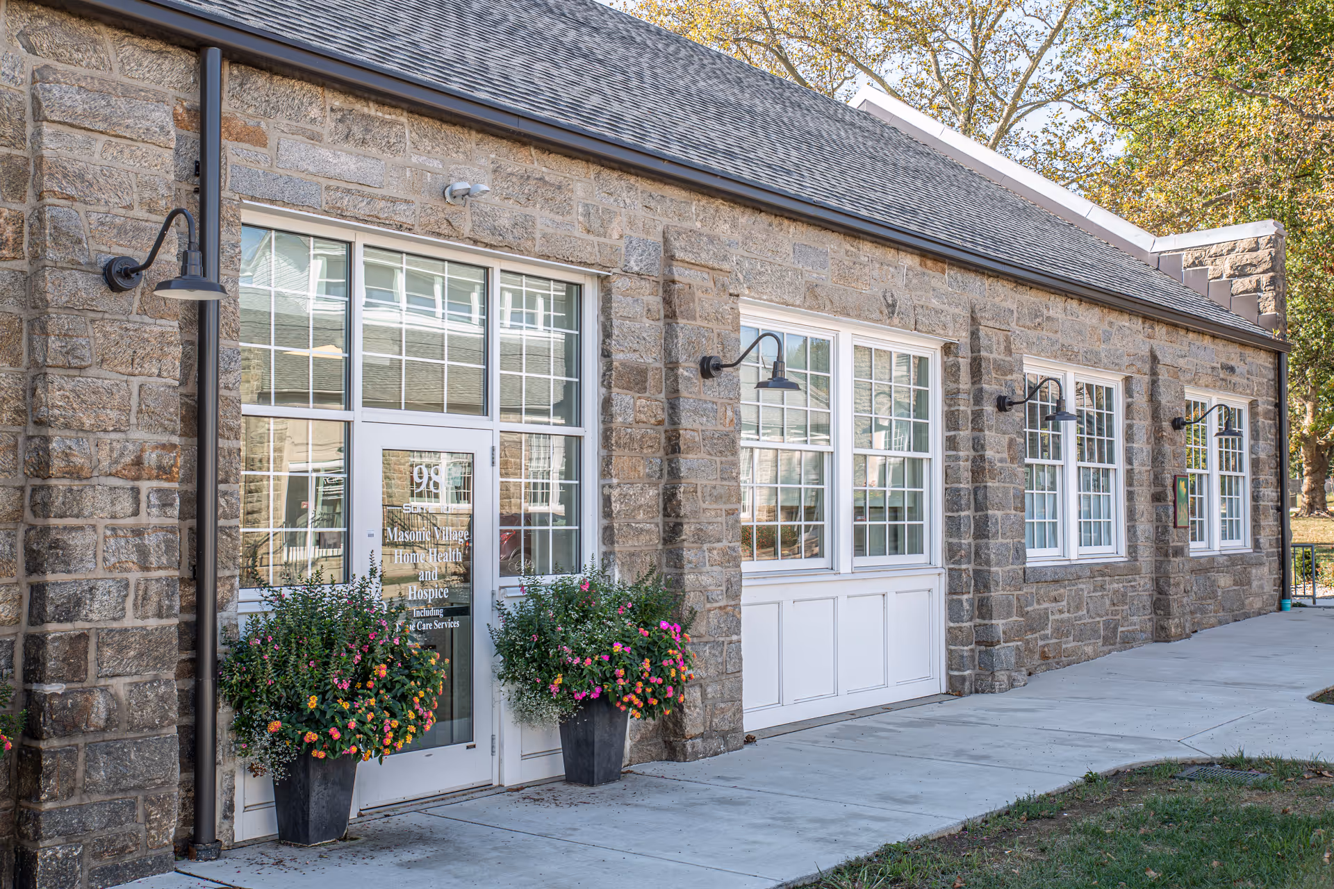 Stone building exterior with large windows, glass doors, and flowering planters at entrance.