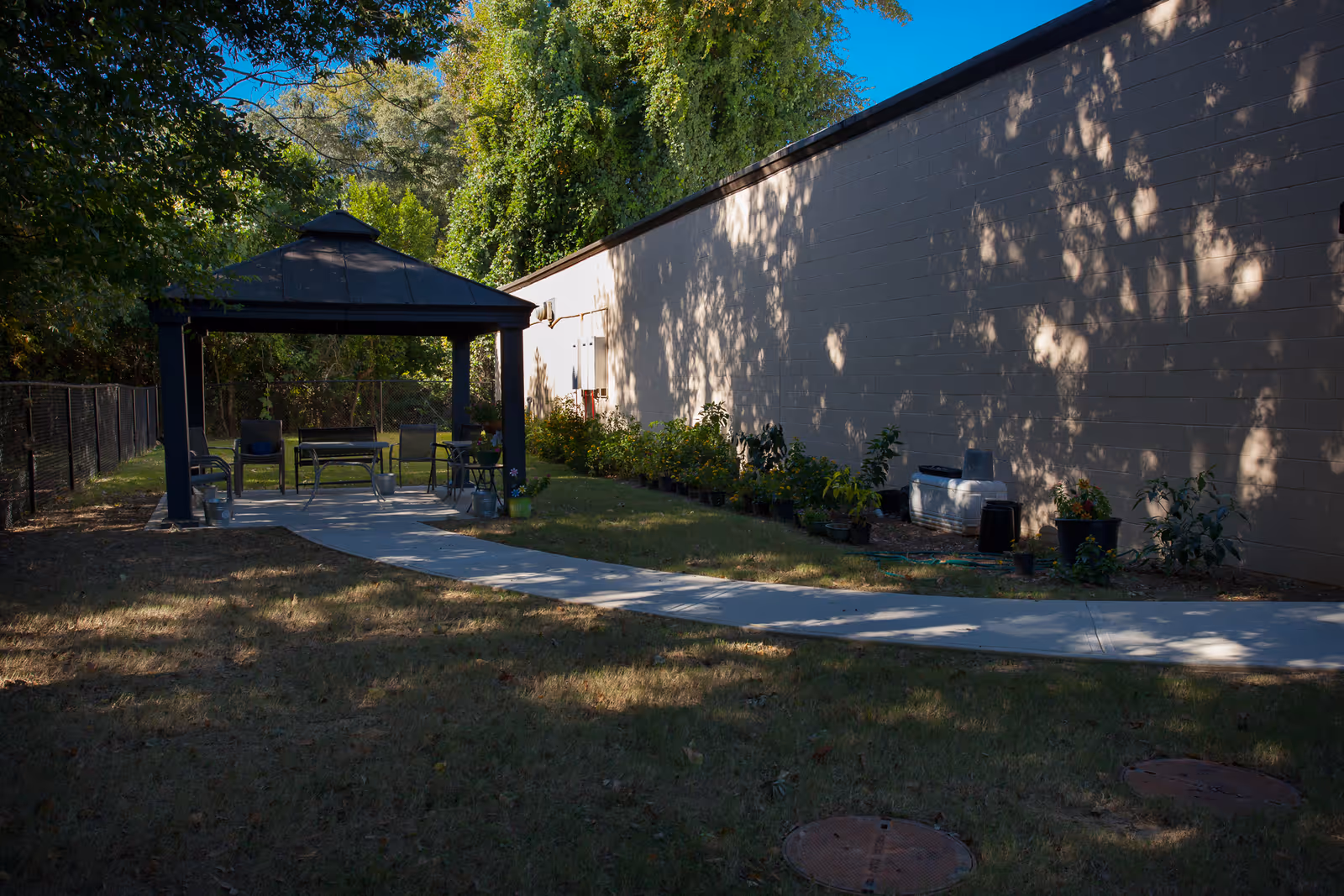 Shaded outdoor patio with a gazebo, tables, chairs, and potted plants beside a building.