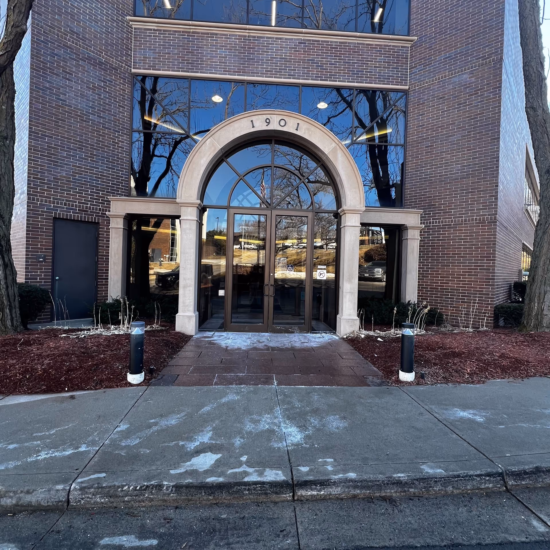 Brick building with a large arched glass entrance and reflections of trees in the windows.