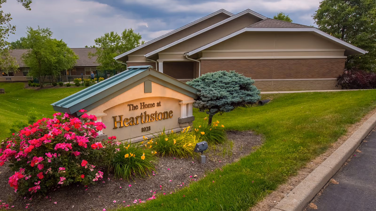 Entrance sign reading "The Home at Hearthstone" surrounded by flowers and landscaping with the facility building in the background.