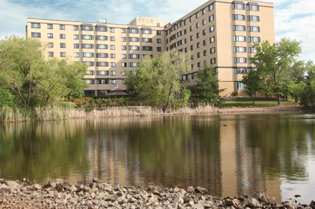 Large beige apartment building beside a pond with trees and rocky shoreline.