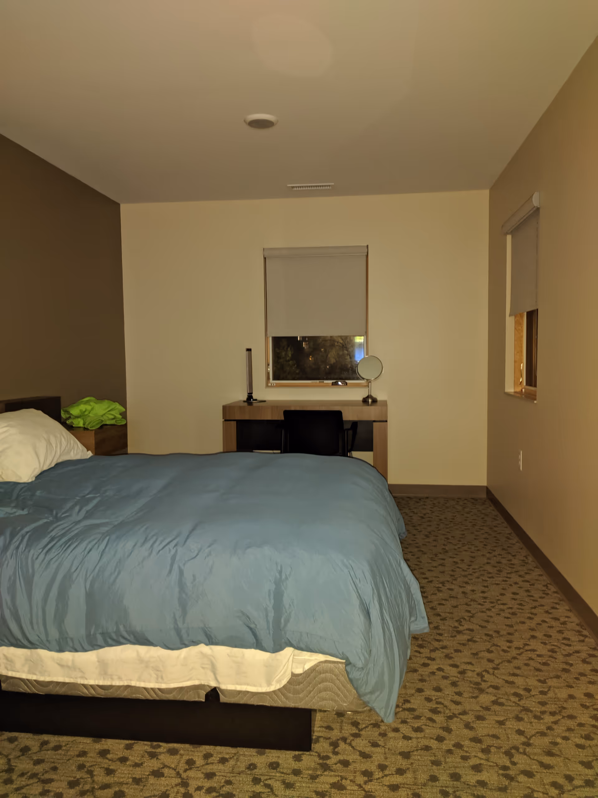 A furnished bedroom in a senior living facility with bed, desk, mirror, and window.