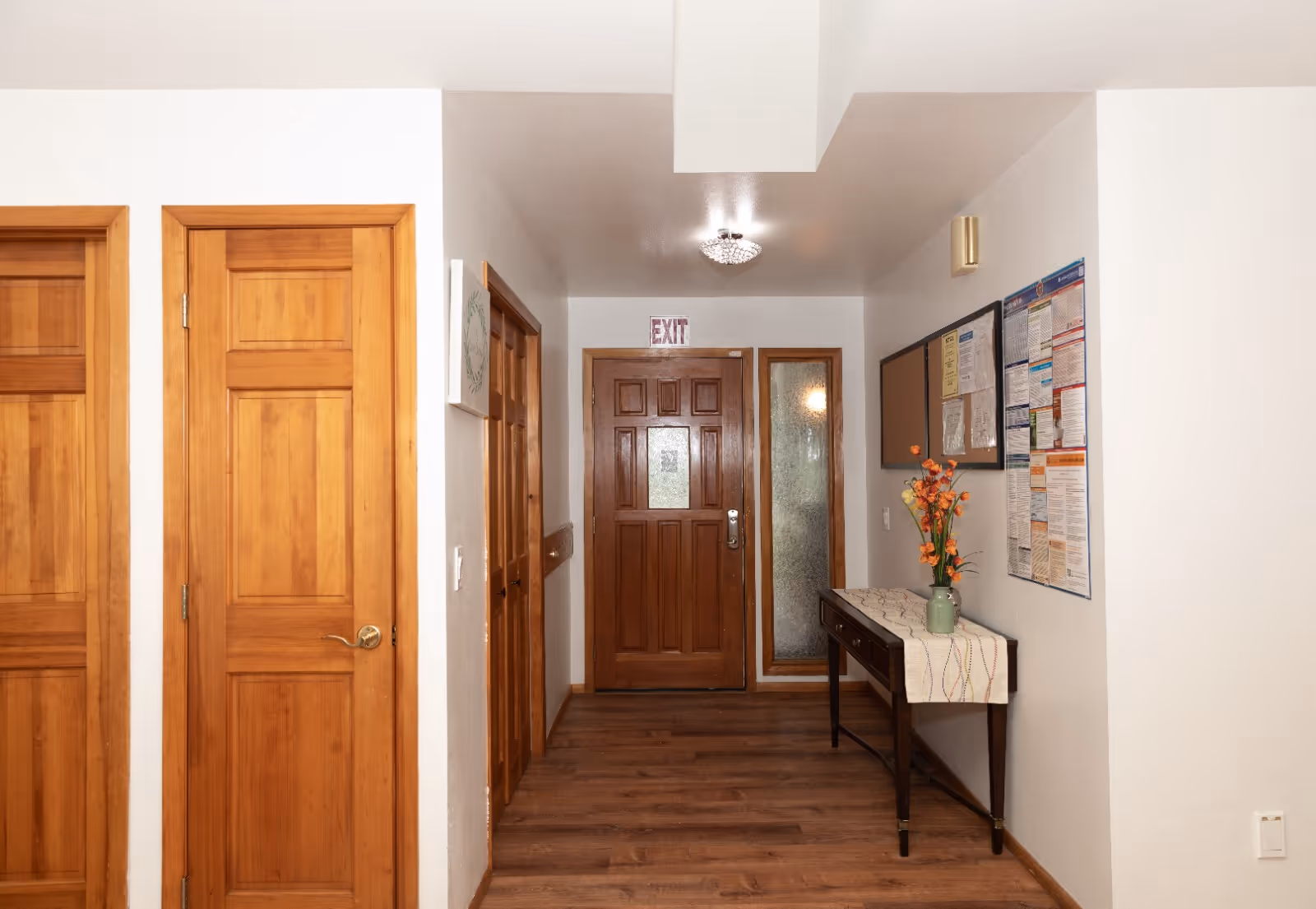 Wooden doors line a bright hallway with a bulletin board and vase on a table.