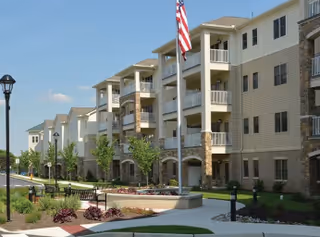 Exterior view of a multi-story senior living facility with balconies, an American flag on a flagpole, landscaped garden beds, benches, and a paved walkway under a clear blue sky.