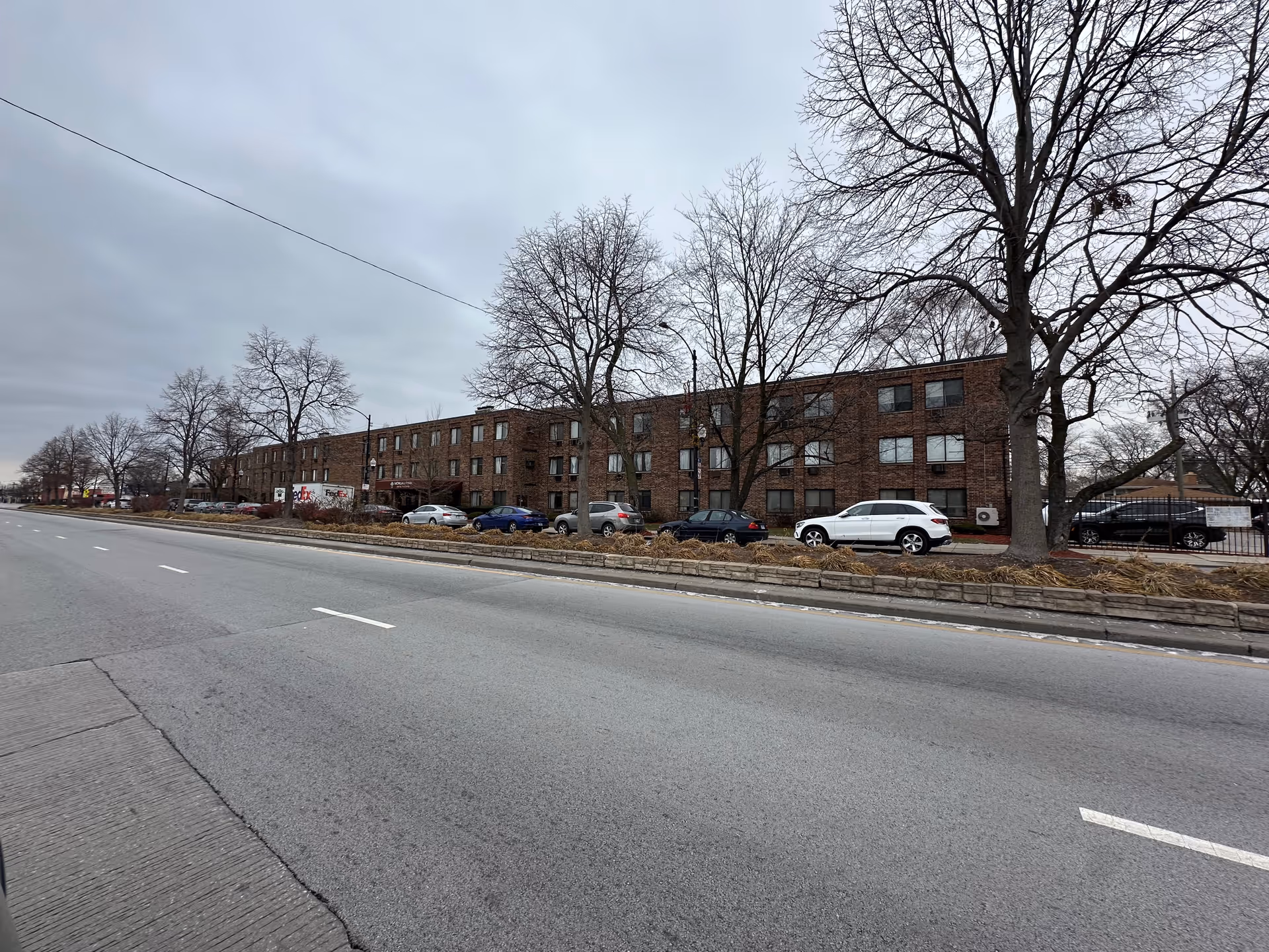 Long three-story brick building with parked cars and bare trees along a wide street under an overcast sky.