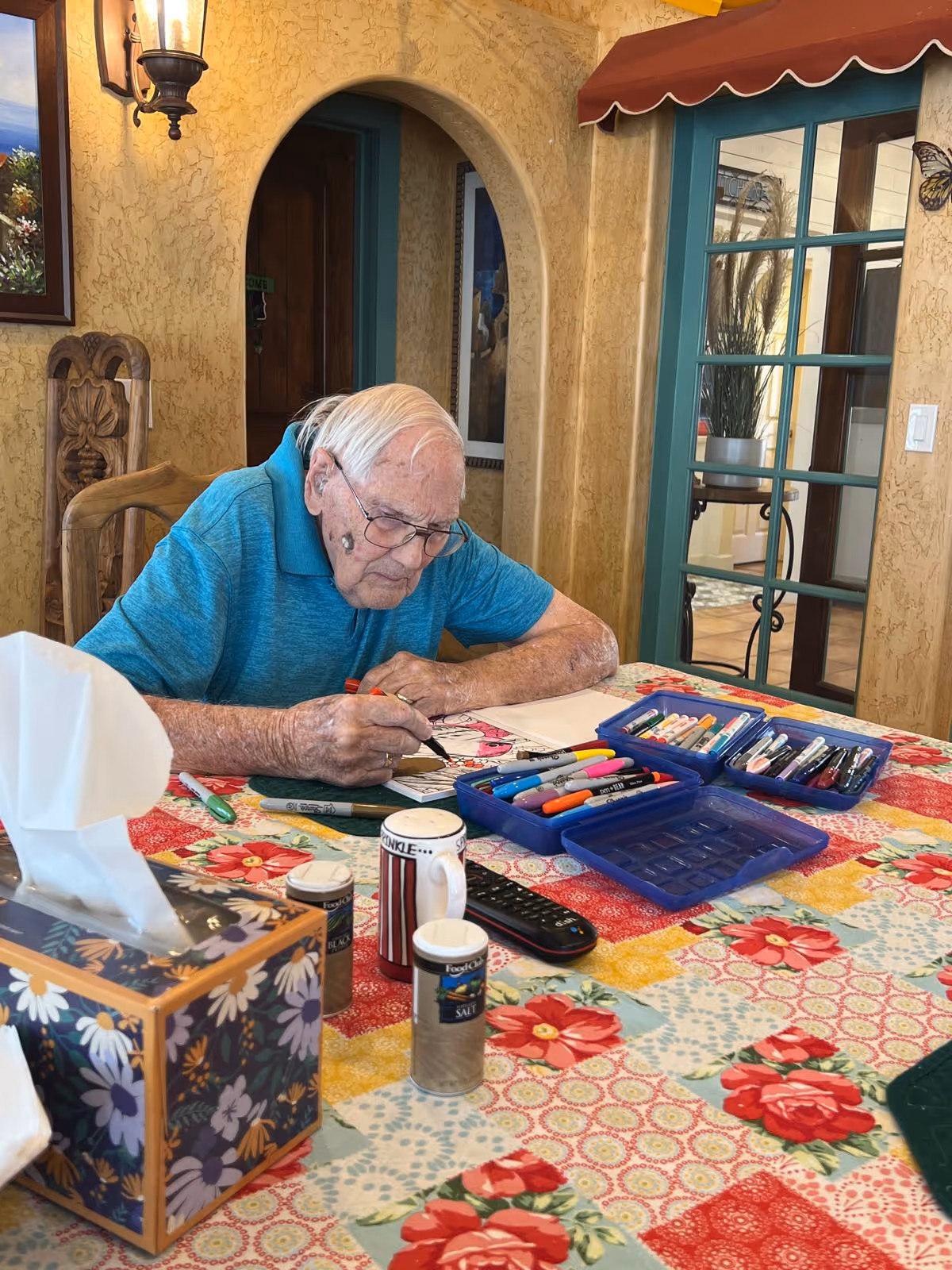 An elderly man with white hair and glasses is sitting at a table covered with a colorful floral tablecloth. He is focused on coloring in a book using various markers and pens spread out in blue containers on the table. The room has textured yellow walls, a wooden chair, and a green-framed glass door with a red awning above it. There is a tissue box, salt and pepper shakers, and a remote control also on the table.