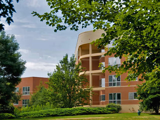 Multi-story brick retirement community building with curved balconies surrounded by trees and a grassy lawn.