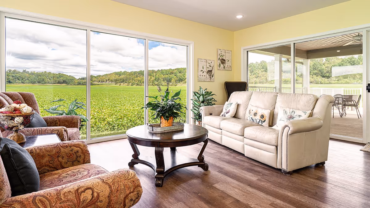 A bright living room with large sliding glass doors offering a view of a green field and trees outside. The room features a beige leather sofa with floral pillows, two patterned armchairs, a round wooden coffee table with a potted plant, and light yellow walls with botanical artwork. Outside on the patio, there is a table with chairs.