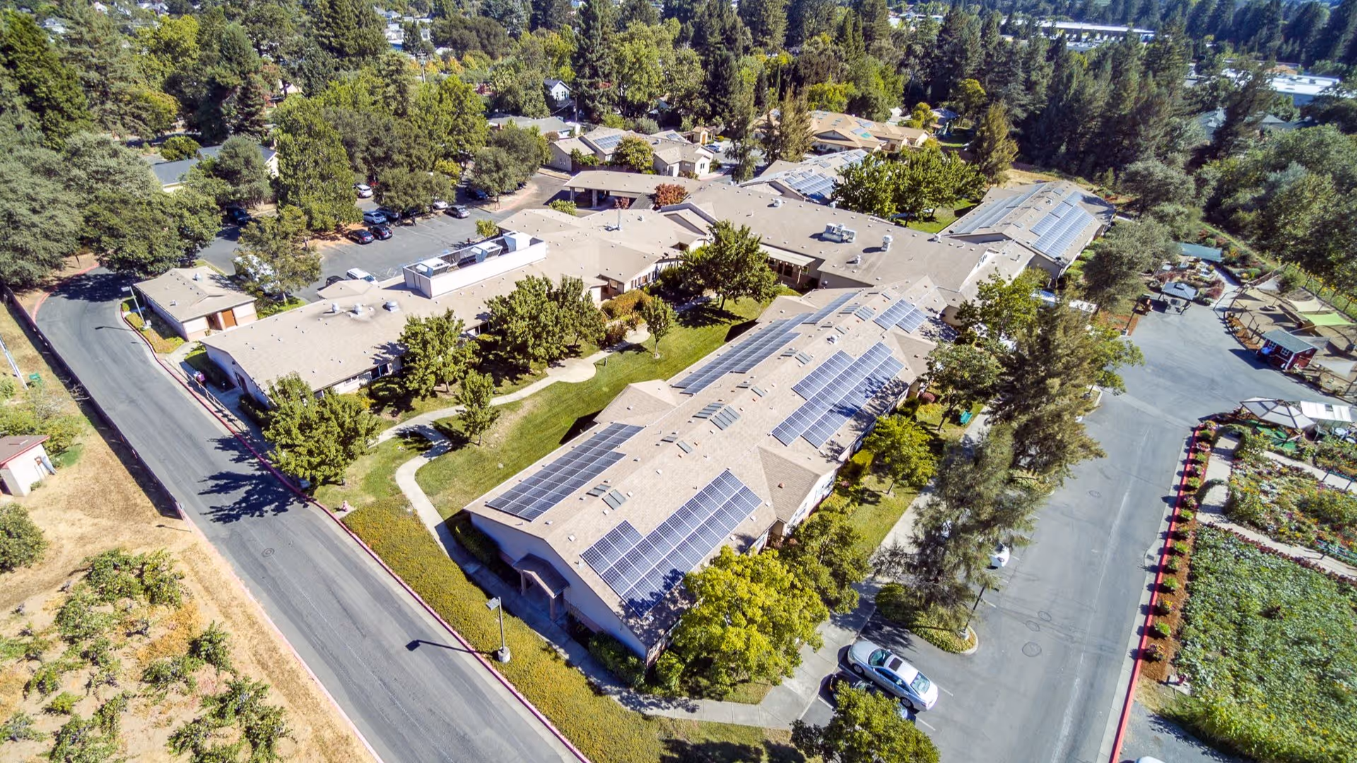 Aerial view of a senior living campus featuring connected buildings with solar panels on the roofs, parking areas and surrounding trees.