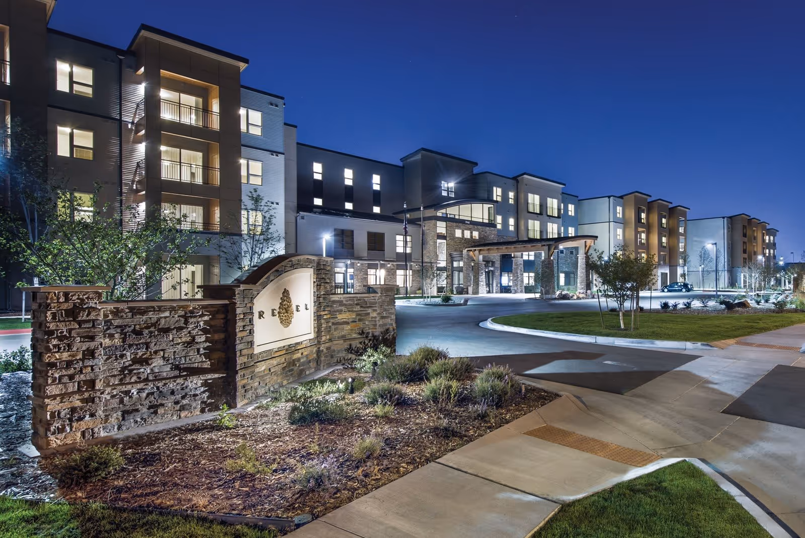Nighttime exterior view of Revel Province, a modern multi-story senior living facility with well-lit windows, a stone entrance sign, landscaped grounds, and a driveway leading to the main entrance.