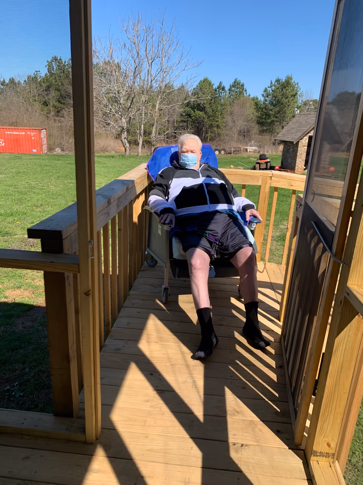 Elderly person in a wheelchair sits on a wooden outdoor porch, wearing a mask.