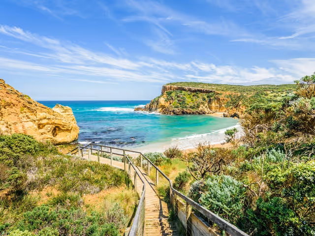 Wooden boardwalk winds through coastal scrub toward turquoise sea and cliffs under clear sky.