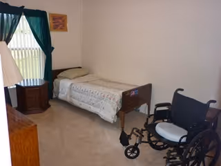 Bedroom in a senior facility with a bed, wheelchair, and window curtains.