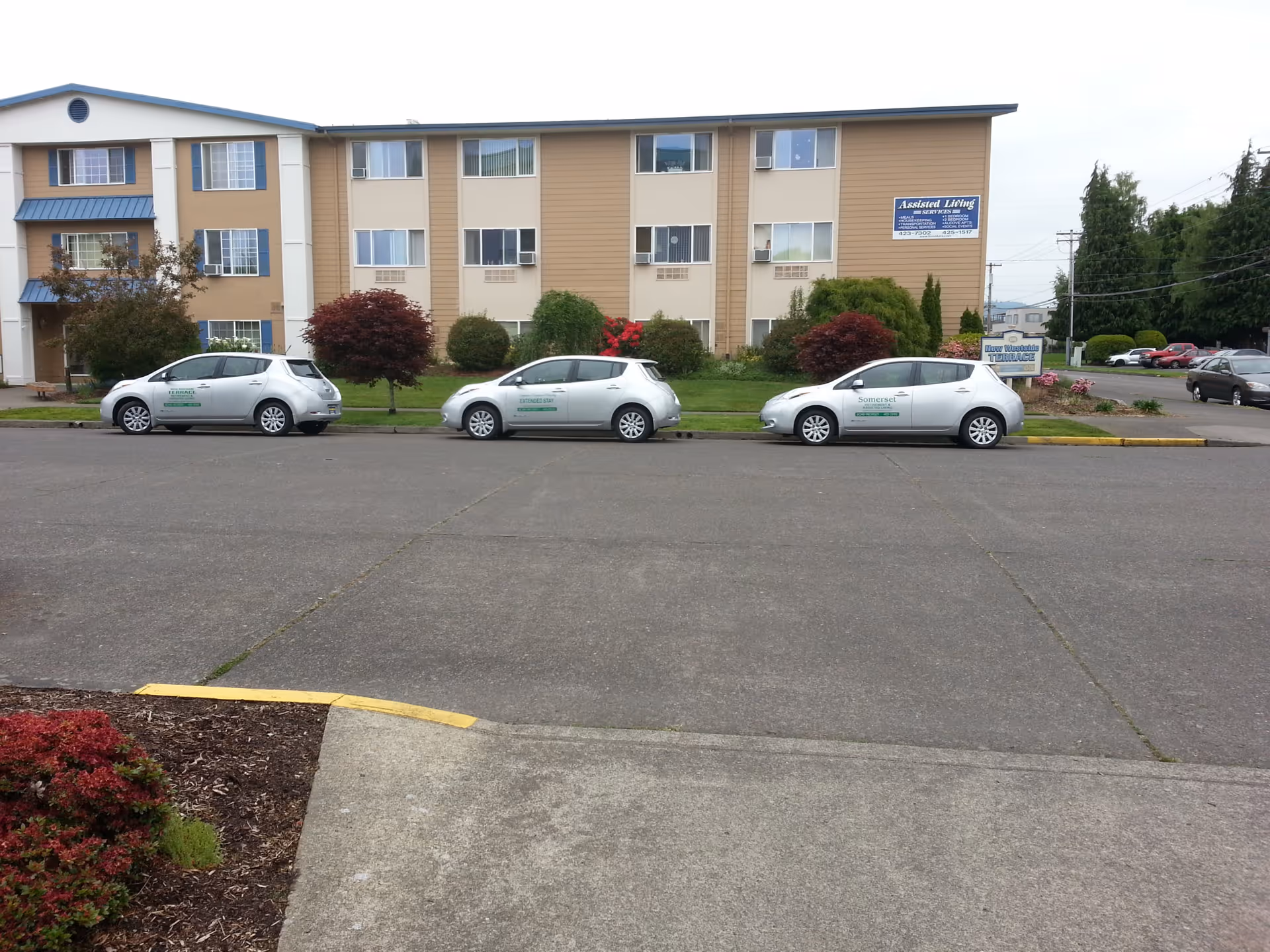 Front view of a three-story assisted living building with parked cars and landscaped grounds.
