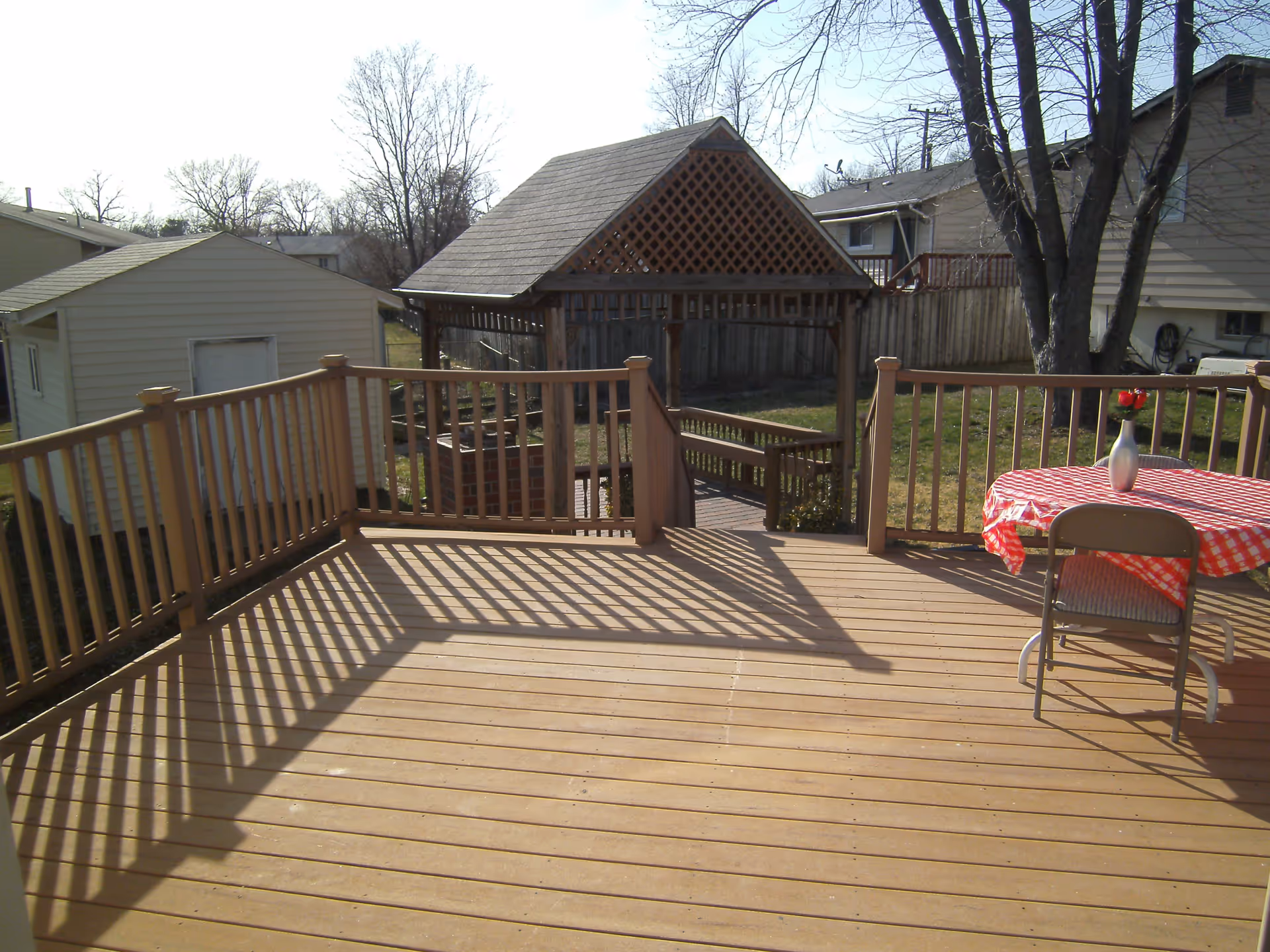 Sunlit wooden backyard deck with railing, a small table covered in a red-checkered cloth and vase, and a wooden gazebo with neighboring houses beyond.